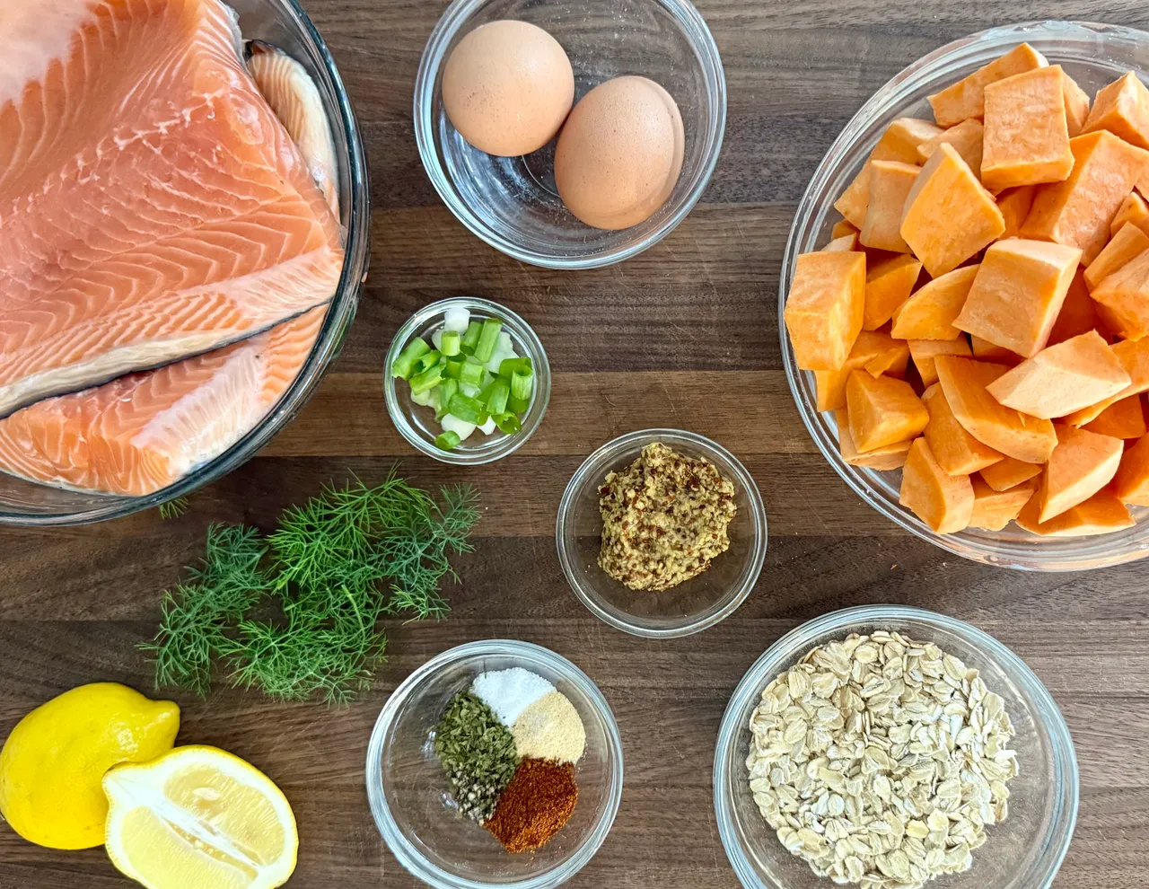 Salmon sweet potato cake ingredients including fresh salmon, cubed sweet potatoes, rolled oats, eggs, Dijon mustard, dill, lemon, green onions, and seasonings before preparation.