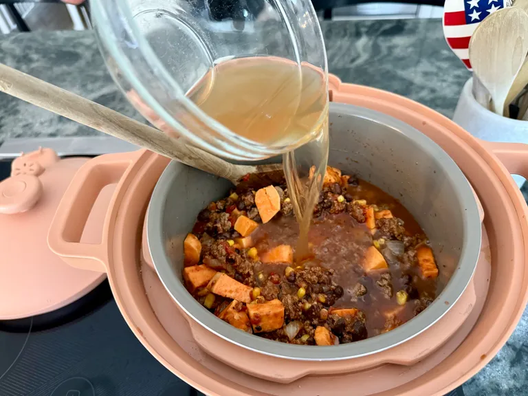 Pouring beef broth into a DreamCooker with ground beef, sweet potatoes, black beans, and vegetables while making healthy beef and sweet potato chili.